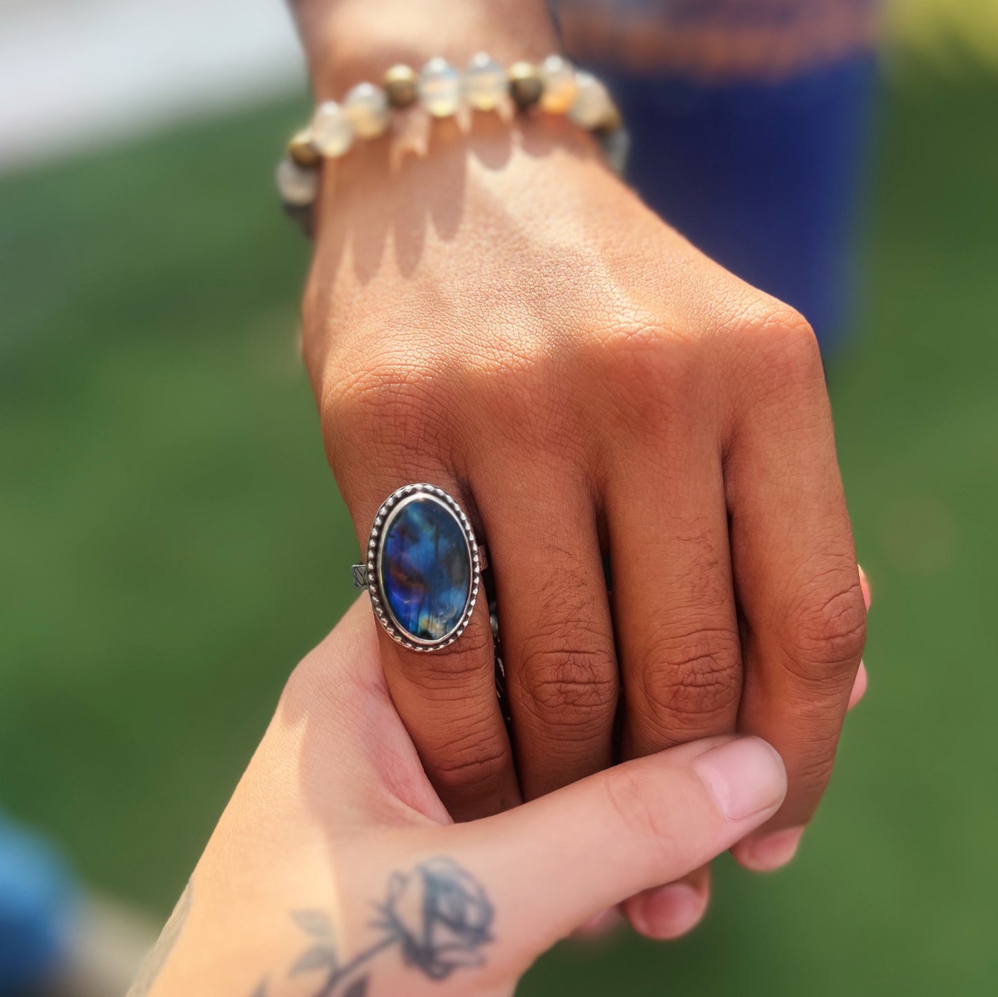 Two hands holding each other with a Labradorite stone set sterling silver handmade ring on one finger, against a blurred green background.